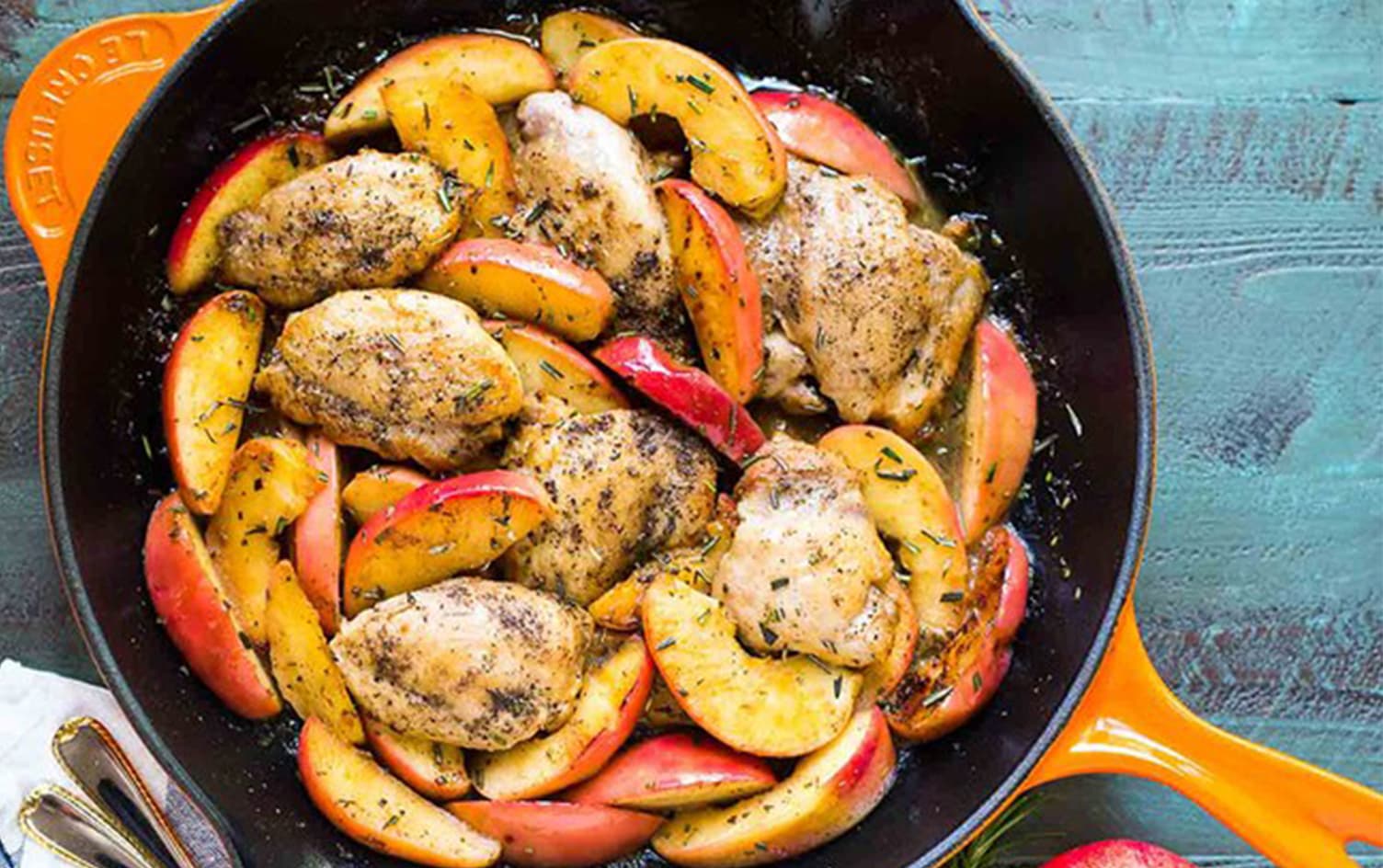 A skillet filled with cooked chicken thighs and sliced apples, perfect for cozy dinners. The dish is seasoned with herbs and appears to be freshly made. The skillet handle is orange, and the background surface is a painted, textured teal color. A portion of a fork and napkin is visible on the side. MyFitnessPal Blog