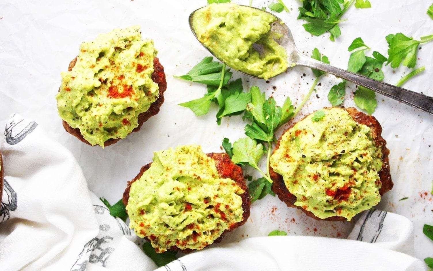 Three juicy burger patties topped with chunky guacamole are arranged on parchment paper, accompanied by a spoonful of guacamole and scattered parsley leaves. Ideal for batch-cooked meals, this arrangement also features a cloth napkin with a charming print design beside the food. MyFitnessPal Blog
