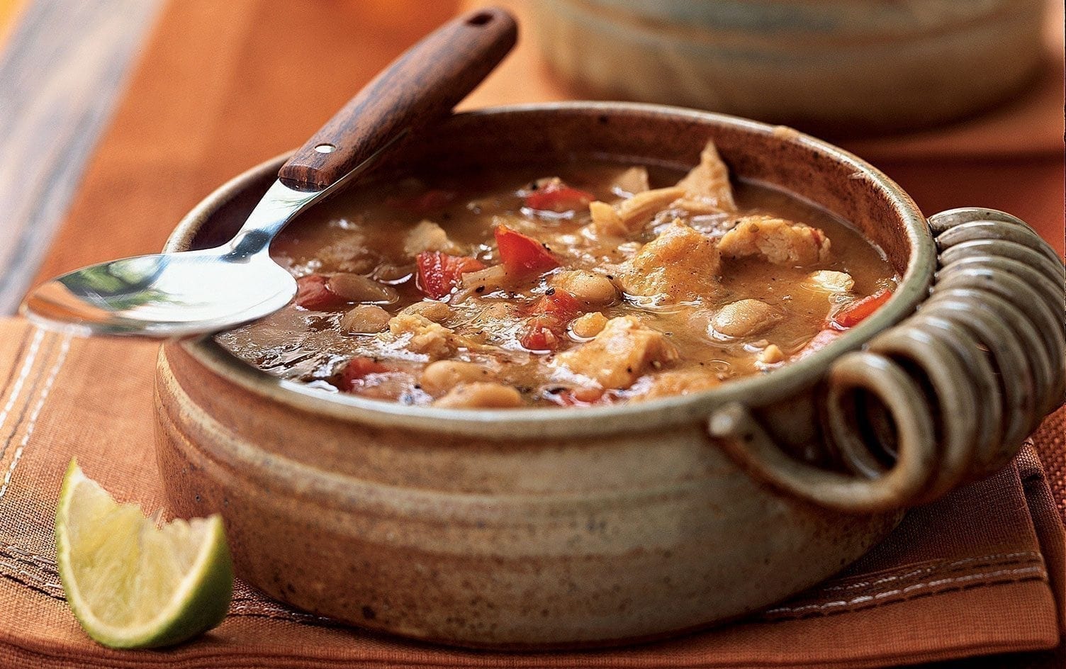 A bowl of vegetable soup containing beans, corn, tomatoes, and other vegetables. The high-fiber recipe is served in a rustic, ceramic bowl with a metal spoon resting on the edge. A lime wedge is placed next to the bowl on a brown placemat. One of 15 recipes guaranteed to leave you satisfied. MyFitnessPal Blog