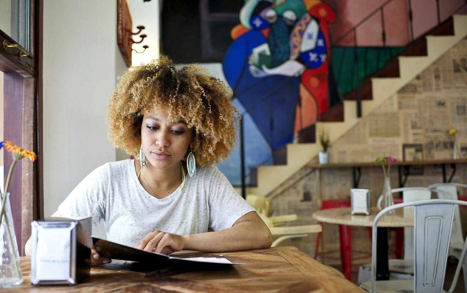 A woman with curly hair sits at a wooden table in a café, reading a vegetarian menu. The café has a vibrant mural on the wall behind her and a staircase decorated with newspaper clippings. She is wearing a white t-shirt and large hoop earrings. MyFitnessPal Blog