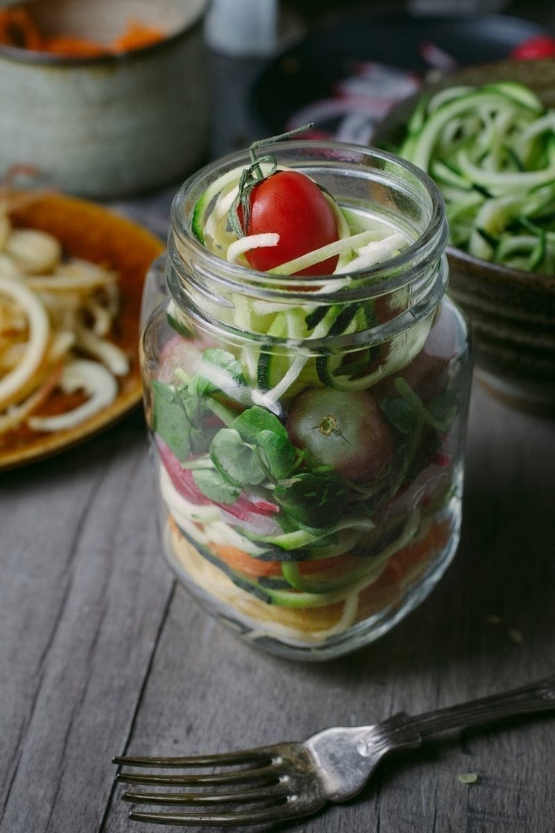 A clear mason jar filled with spiralized vegetables and a small cherry tomato on top. The jar is placed on a wooden table next to a fork. Additional spiralized vegetables are in the background on various plates, showcasing the perfect arrangement for making refreshing mason jar salads. MyFitnessPal Blog