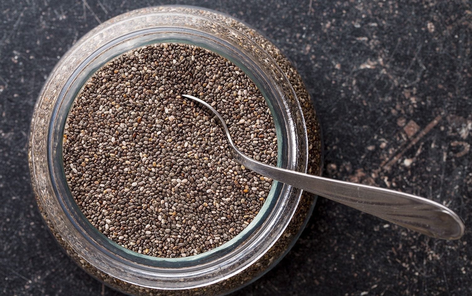 A close-up view of an open jar filled with chia seeds. A small metal spoon is placed inside the jar. The background features a textured, dark surface, highlighting the tiny details of the nutrient-packed chia seeds. MyFitnessPal Blog