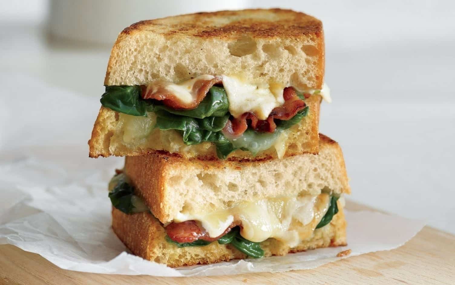 A close-up of halved sandwiches with toasted bread, spinach, melted cheese, and pieces of bacon. The sandwich halves are stacked on top of each other, resting on a wooden surface with a piece of parchment paper. MyFitnessPal Blog