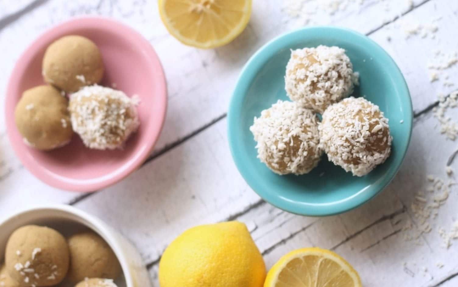 Three bowls containing round, yellowish treats are displayed on a white wooden surface. Two whole lemons and one sliced lemon are placed around the bowls. The mini-breakfasts in the blue bowl are covered in shredded coconut, while the others are plain. MyFitnessPal Blog