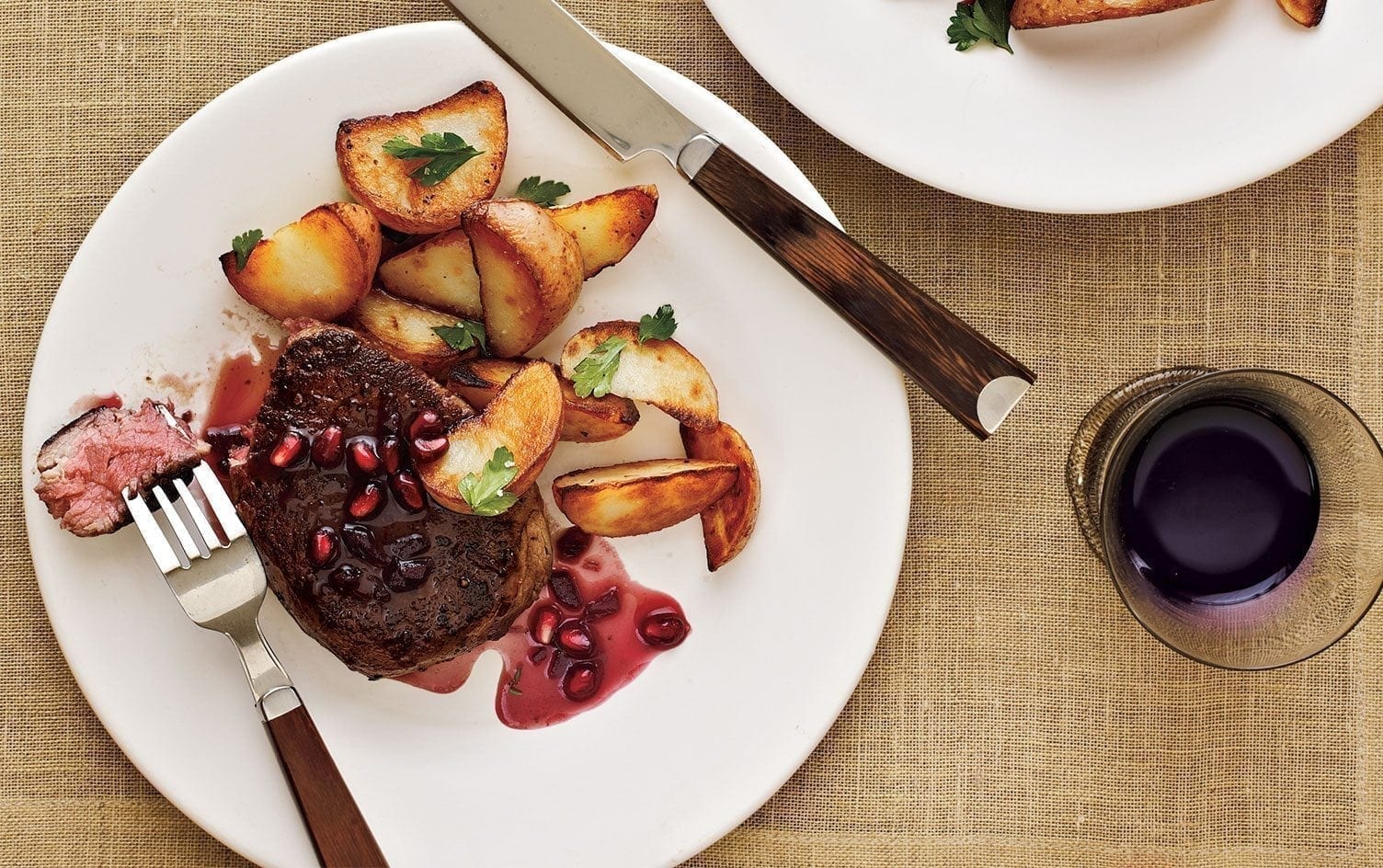 A plate of steak topped with pomegranate seeds and sauce, served with roasted potatoes and garnished with parsley. A knife and fork are placed on the side of the plate. A glass of red wine sits next to the plate on a beige tablecloth, perfect for romantic meals. MyFitnessPal Blog