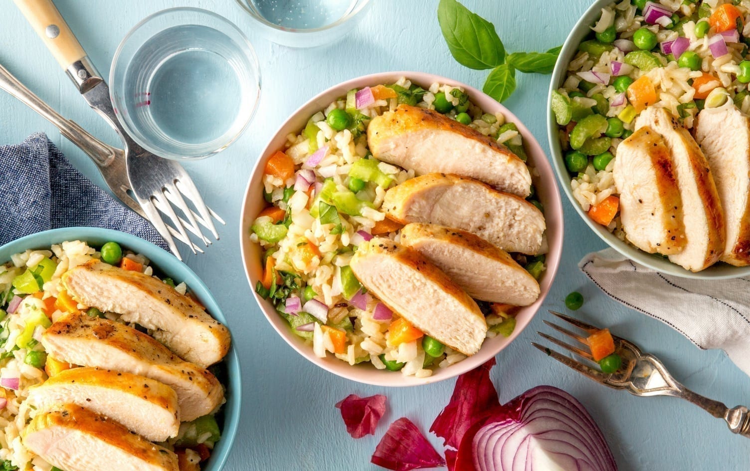Three bowls of grilled chicken slices on rice mixed with vegetables are placed on a light blue table. Two forks lie beside the bowls, and two glasses of water can be seen. Fresh basil leaves and cut red onion pieces add a burst of color to the scene. MyFitnessPal Blog