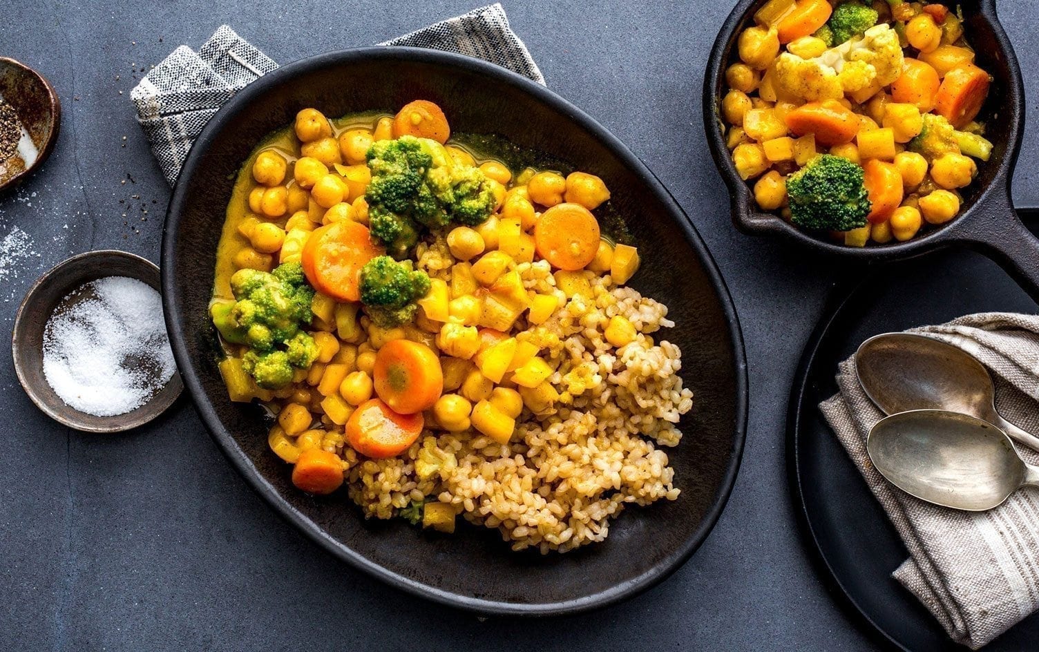 A black oval plate holds a serving of chickpeas, broccoli, and carrots in a yellow sauce alongside a portion of brown rice. Next to it, a bowl with more chickpea curry sits on a striped napkin. Spoons and a small dish of salt are also visible.—a perfect chickpea recipe for any dinner table. MyFitnessPal Blog