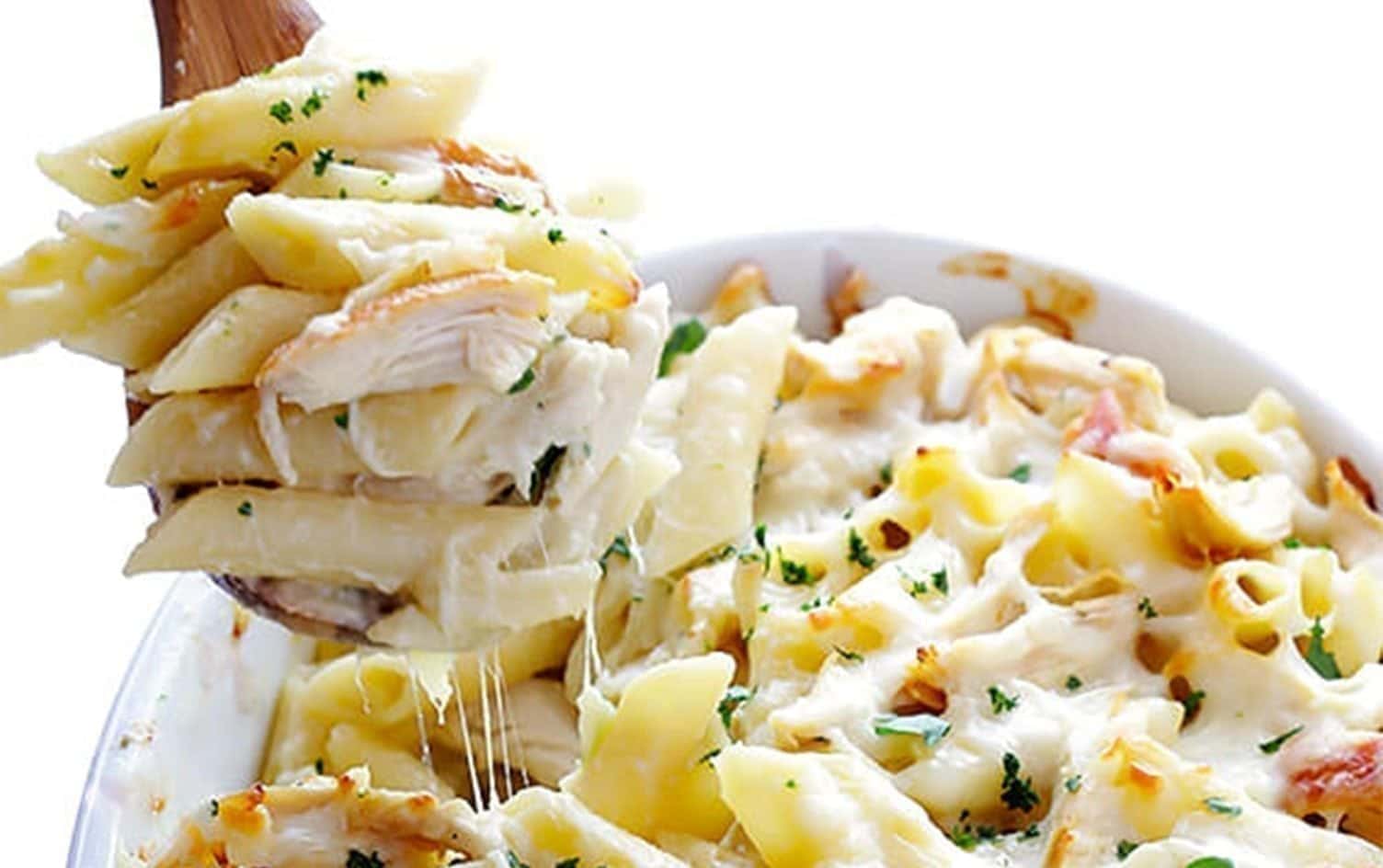 A wooden spoon scoops up creamy baked pasta from a white casserole dish. The pasta is topped with melted cheese and garnished with chopped parsley. Shredded pieces of chicken are mixed into the dish, showcasing one of the tastiest chicken casseroles as the cheese stretches while lifting the pasta. MyFitnessPal Blog