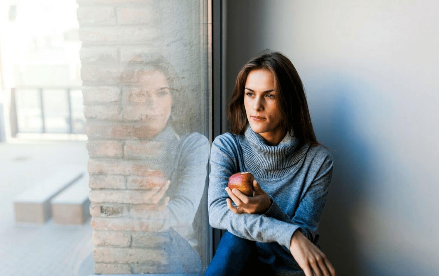 A woman with long brown hair and wearing a gray turtleneck sweater sits by a window holding an apple, contemplating her intermittent fasting routine. She looks outside, with her reflection visible on the glass. The background includes a brick wall and out-of-focus outdoor scenery. MyFitnessPal Blog