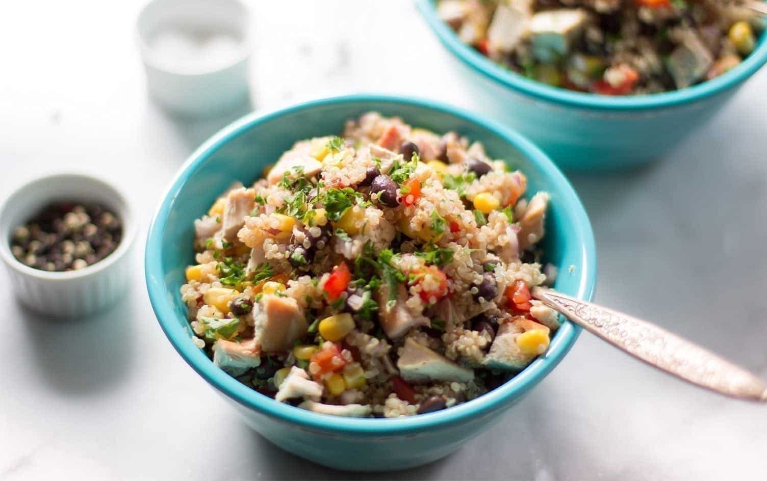 A close-up image of a bowl of quinoa and chicken salad topped with chopped vegetables such as red bell peppers, corn, black beans, and herbs. The salad is served in a turquoise bowl with a spoon inside. Another bowl of salad and some small condiment bowls are in the background. MyFitnessPal Blog