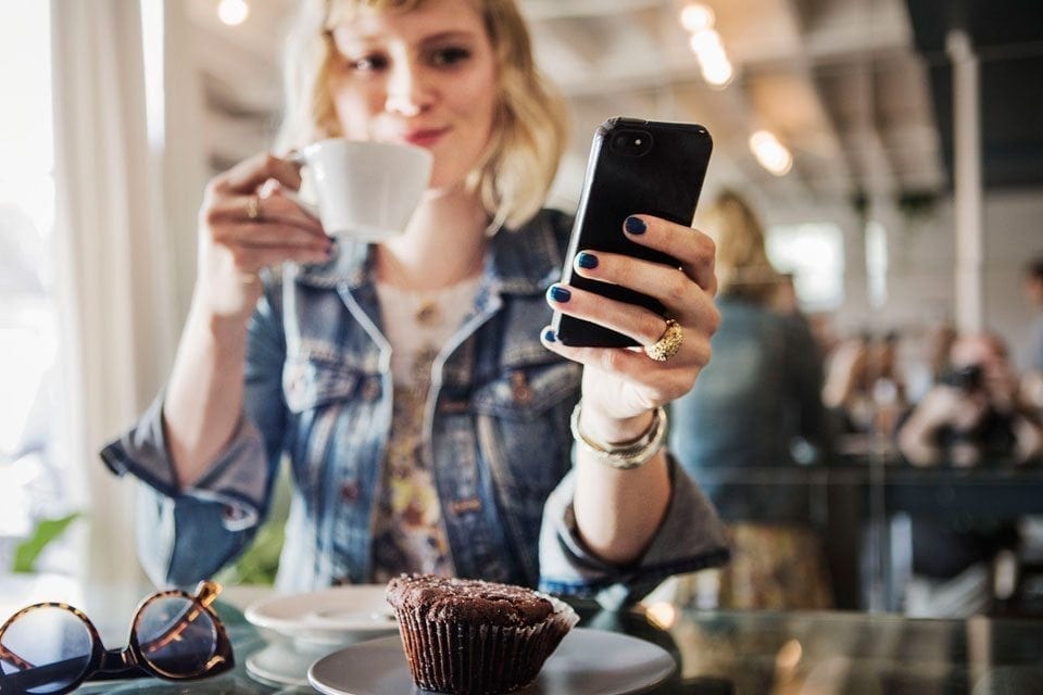 A person with blond hair holds a white cup in one hand and a smartphone running MyFitnessPal in the other while seated at a table. A chocolate muffin on a plate and a pair of glasses are on the table. The person appears relaxed, enjoying their drink and checking their phone. MyFitnessPal Blog