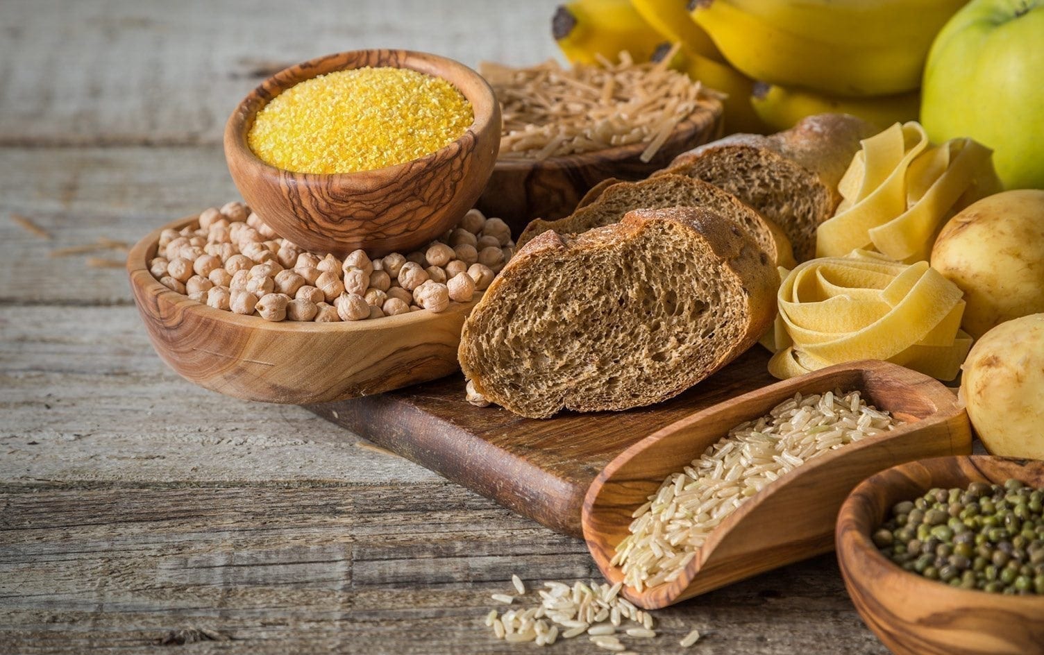 A wooden table displays a variety of foods rich in carbohydrates, including chickpeas, cornbread, spaghetti, sliced whole wheat bread, pappardelle pasta, rice, bananas, and apples. Some foods are in small wooden bowls or scattered on a wooden board. MyFitnessPal Blog