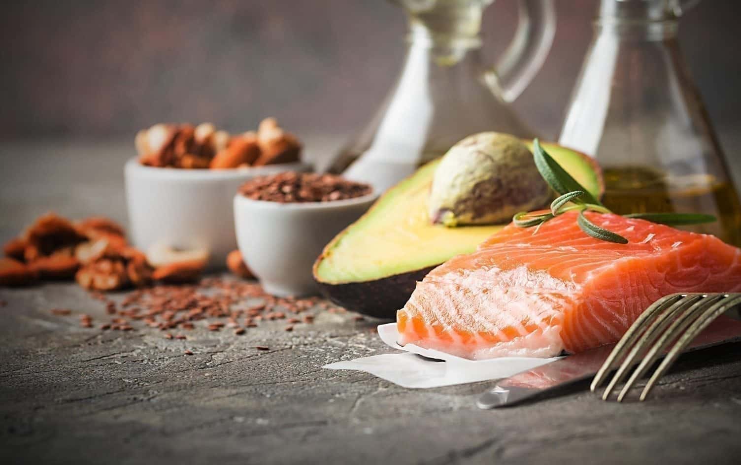 A piece of raw salmon fillet is placed on parchment paper next to a fork, with a halved avocado, bowls of nuts and seeds, and two bottles of oil in the background. The items are arranged on a grey textured surface. MyFitnessPal Blog