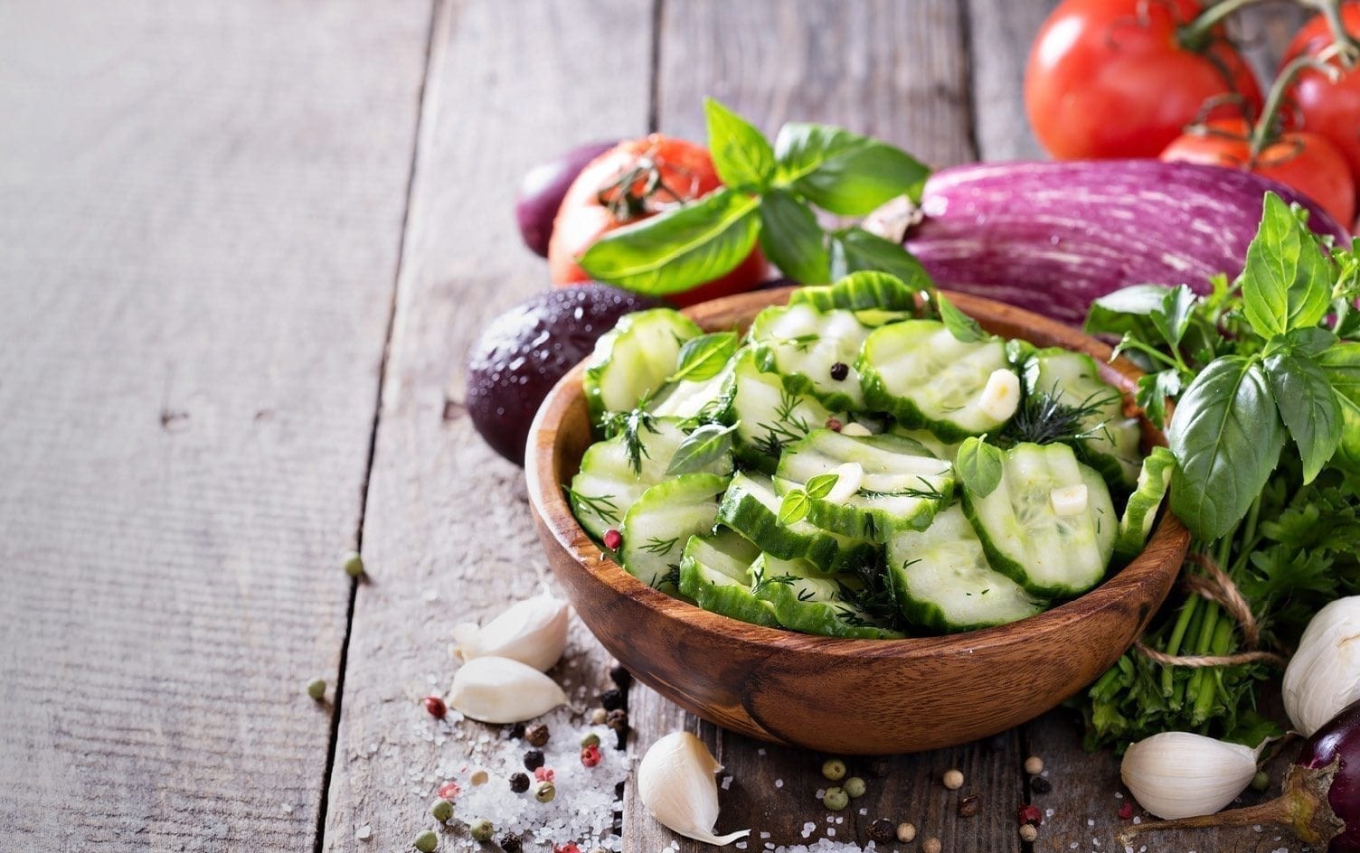 A wooden bowl filled with cucumber slices garnished with fresh herbs. Surrounding the bowl are various vegetables, including tomatoes, eggplants, garlic cloves, and avocado, all arranged on a rustic wooden surface. MyFitnessPal Blog