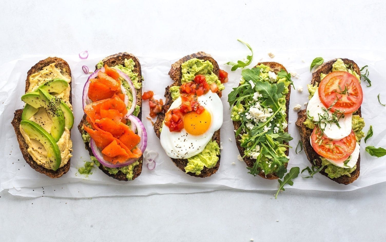 Five slices of avocado toast topped with a variety of ingredients are arranged in a row. Toppings include hummus, smoked salmon, sliced onions, a fried egg, salsa, arugula, tomatoes, and cheese. The toast is placed on parchment paper on a light surface. MyFitnessPal Blog