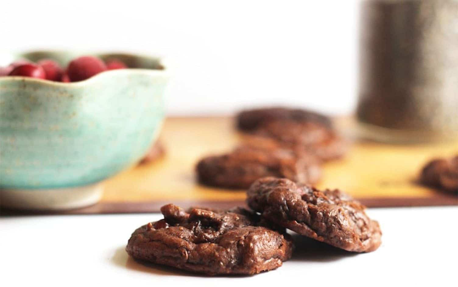 Two vegan chocolate cookies are in focus on a white surface, with a blurred green bowl filled with red fruits and more out-of-focus cookies in the background on a wooden surface. A jar is also partially visible in the background, perfect for those exploring vegan chocolate dessert recipes. MyFitnessPal Blog