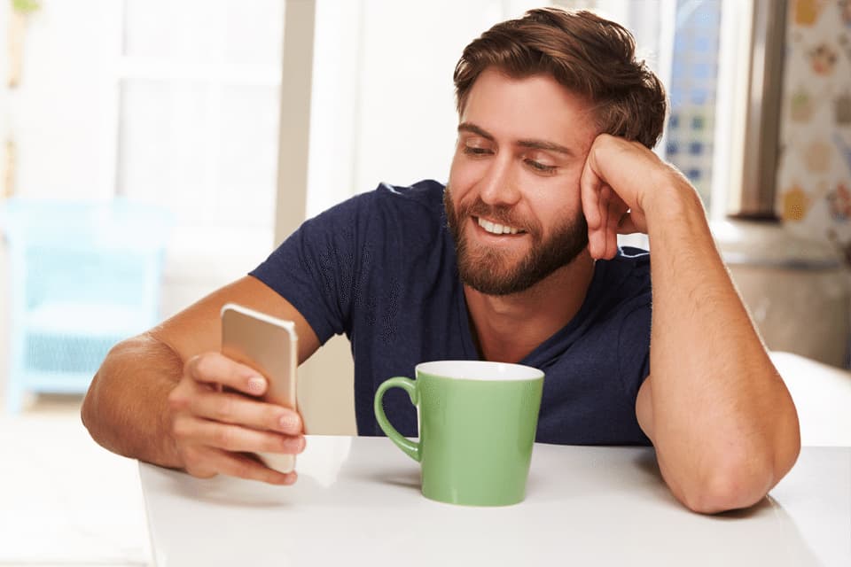A man with brown hair and a beard is sitting at a table, holding a smartphone in his right hand and smiling while looking at health goals on the screen. His left elbow rests on the table, supporting his head with his hand. A large green mug is placed on the table. MyFitnessPal Blog